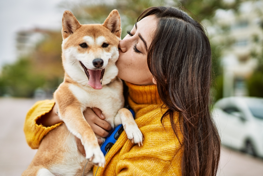 Smiling woman in a yellow sweater holding and kissing a happy Shiba Inu dog on the street, the dog’s mouth open in a joyful expression - Scottsdale Emergency Vet